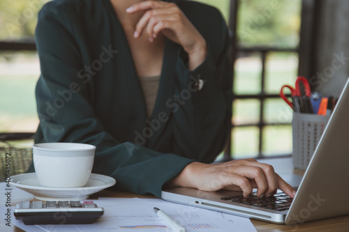 Businesswoman working on laptop in her workstation.Black blank screen smartphone, potted plant, pencil, notes, earphone on wooden desk. Close up
