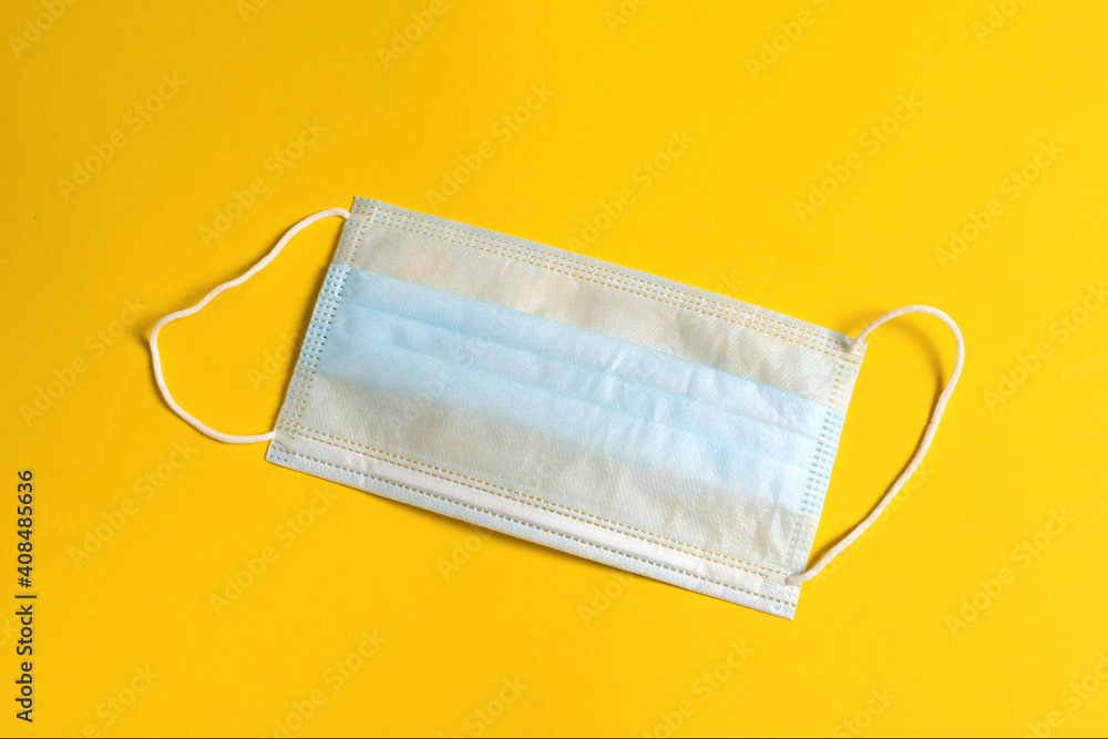 Surgical three-layer mask with rubber ear straps to protect against bacteria. Disposable medical mask covering mouth and nose on yellow background Surgical three-layer mask with rubber ear straps to protect against bacteria. Disposable medical mask covering mouth and nose on yellow background