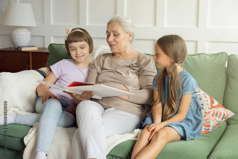 Smiling grandmother and little granddaughters sit on sofa at home reading mockup book together. Gray haired granny or nanny enjoy fairytale with small girls. Education concept