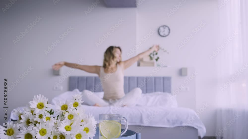 Chamomile flowers at water glass with lemon slice against sleepy young woman waking on bed with blanket and stretching arms and body in early morning Chamomile flowers at water glass with lemon slice against sleepy young woman waking on bed with blanket and stretching arms and body in early morning