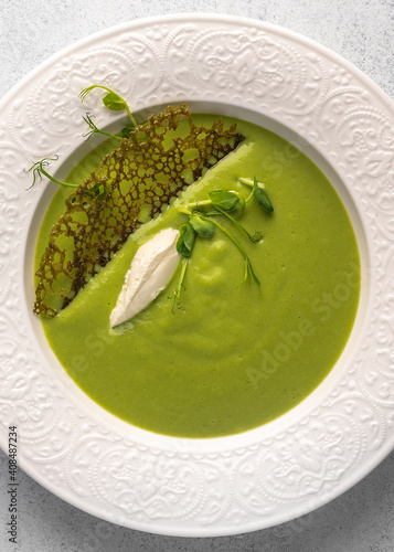 Vertical view homemade soup puree of green peas and coconut milk with tuil and cheese quenelle in white plate on light background, close up photo