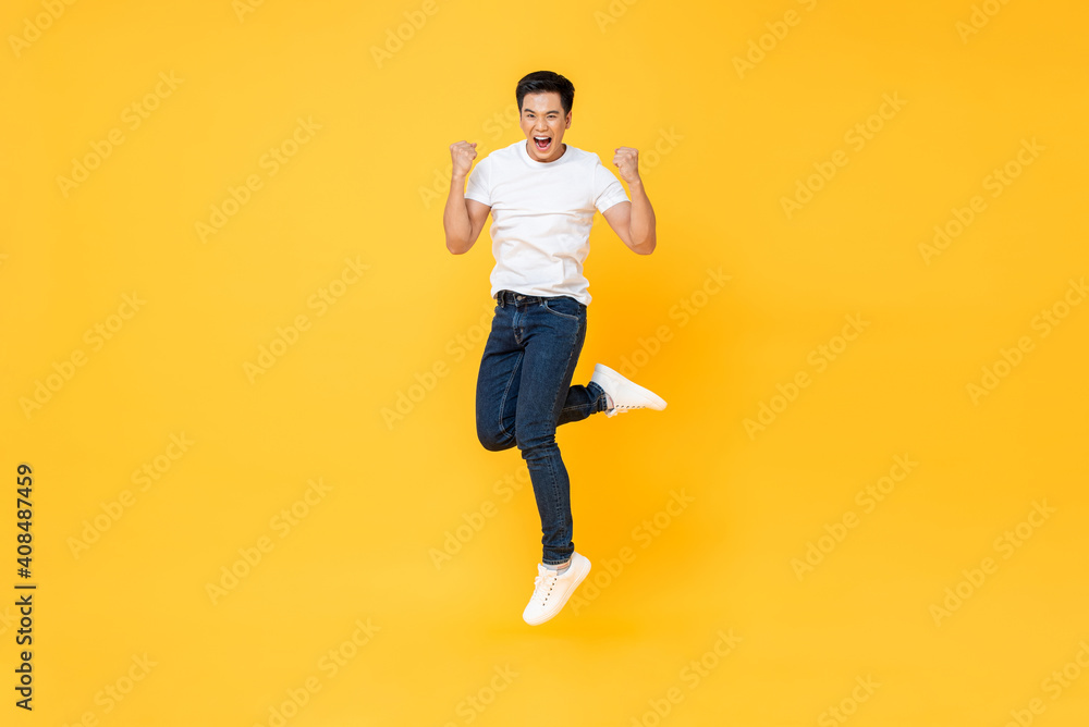 Happy energetic young Asian man jumping yelling and clenching fists isolated on yellow studio background