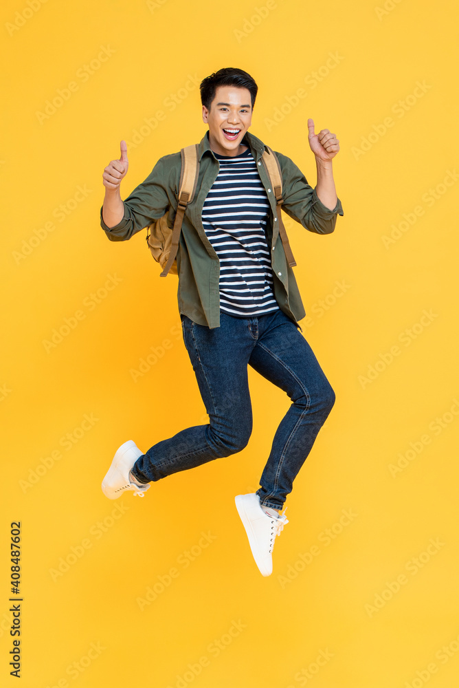 Happy smiling young Asian tourist man with backpack jumping and giving thumbs up isolated on yellow background