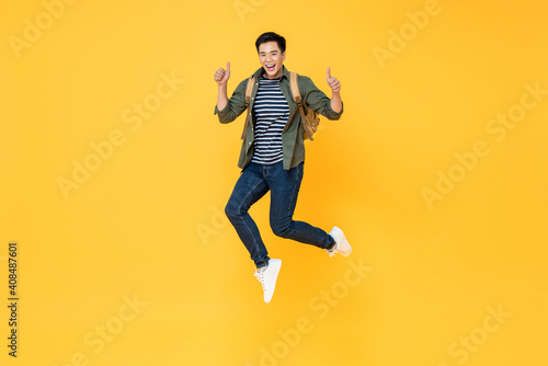 Happy smiling young Asian tourist man with backpack jumping and giving thumbs up isolated on yellow background