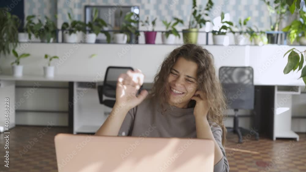 Businesswoman with long loose curly hair talks on video call looking into laptop display and holds smartphone against empty workplaces with pot plants Businesswoman with long loose curly hair talks on video call looking into laptop display and holds smartphone against empty workplaces with pot plants