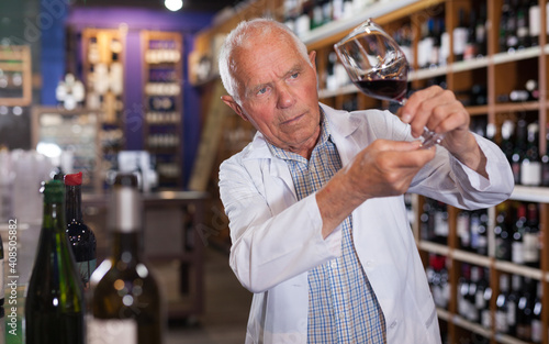 Older man wine producer inspecting quality of red wine, standing in wineshop on background with shelves of wine bottles