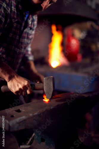 Male Blacksmith Hammering Metalwork On Anvil With Blazing Forge In Background