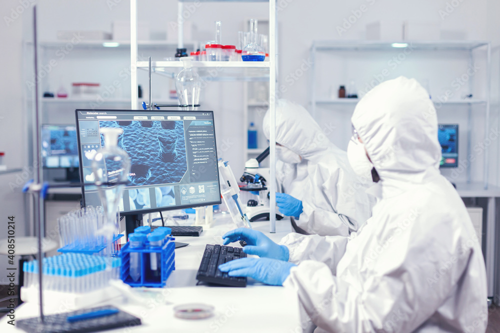 Chemist in ppe suit typing on keyboard checking virus development in equipped laboratory. Medical engineer using computer during global pandemic with coronavirus dressed in coverall. Chemist in ppe suit typing on keyboard checking virus development in equipped laboratory. Medical engineer using computer during global pandemic with coronavirus dressed in coverall.