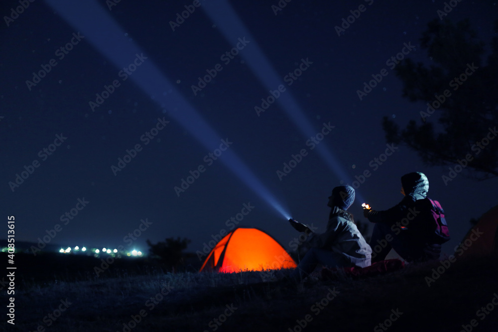 Couple with flashlights near camping tent outdoors at night - obrazy, fototapety, plakaty 