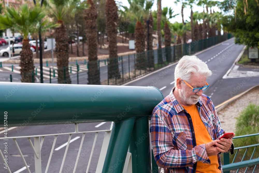 A white-haired senior man standing on an elevated city street with cellphone in the hand reading a message. Old people using modern technology. Boulevard behind him with palm trees