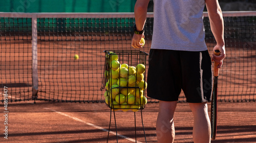 Tennis coach conducts training on red clay court, basket with tennis balls near him. Blurred background. Sports activity and leisure concept. Lifestyle © Elena