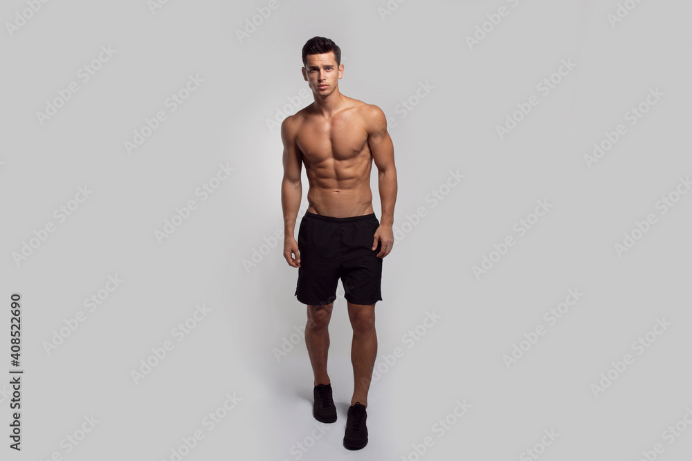 Full length image of a confident young man shirtless torso showing six pack abs, isolated grey background.