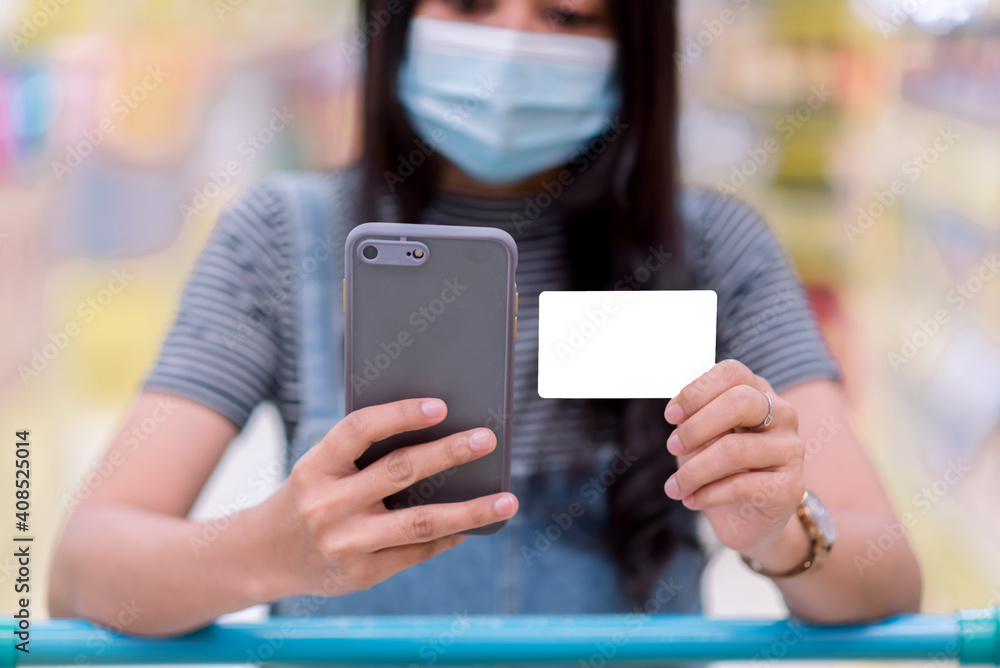 A masked woman is using a smartphone and in her hand is a white credit card. At the supermarket during the COVID-19 pandemic