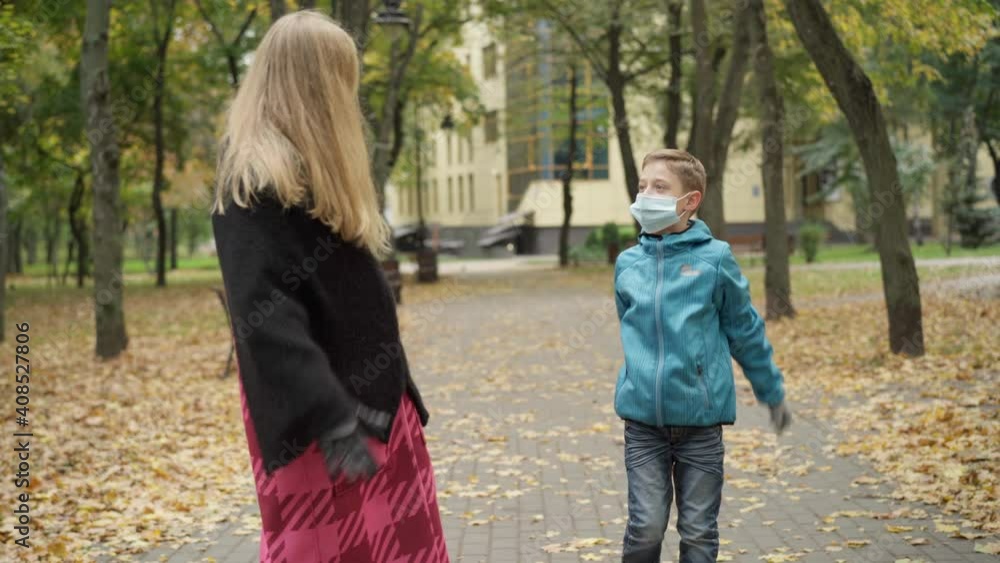 Portrait of excited Caucasian boy in Covid-19 face mask talking with beautiful woman walking in autumn park. Relaxed son strolling with mother outdoors on coronavirus pandemic. New normal. Portrait of excited Caucasian boy in Covid-19 face mask talking with beautiful woman walking in autumn park. Relaxed son strolling with mother outdoors on coronavirus pandemic. New normal.