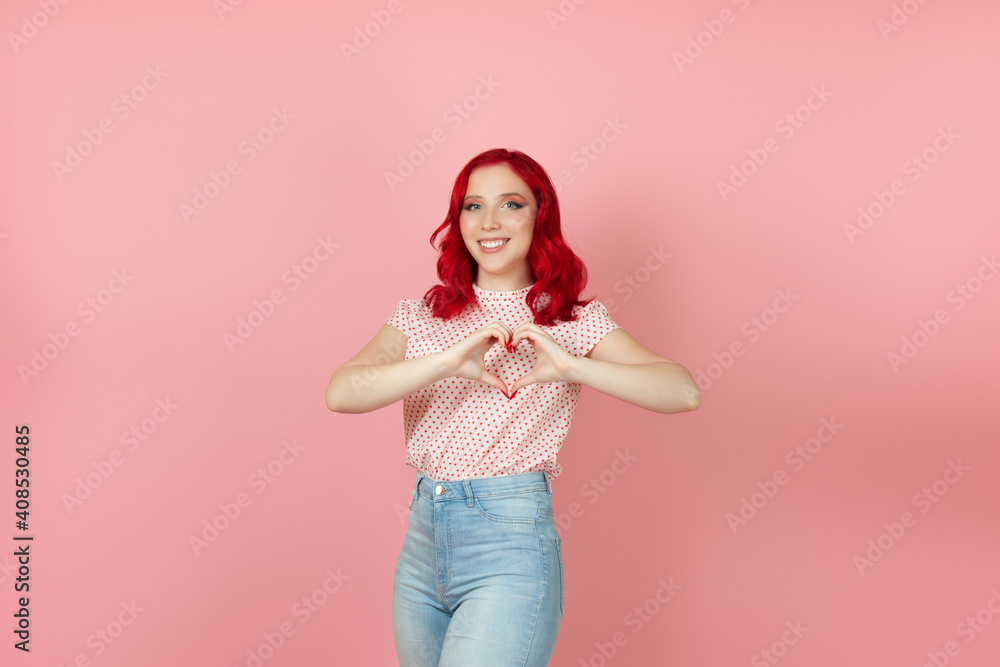 a smiling, contented woman with red hair makes a heart sign from her fingers at her chest , isolated on a pink background. a smiling, contented woman with red hair makes a heart sign from her fingers at her chest , isolated on a pink background.