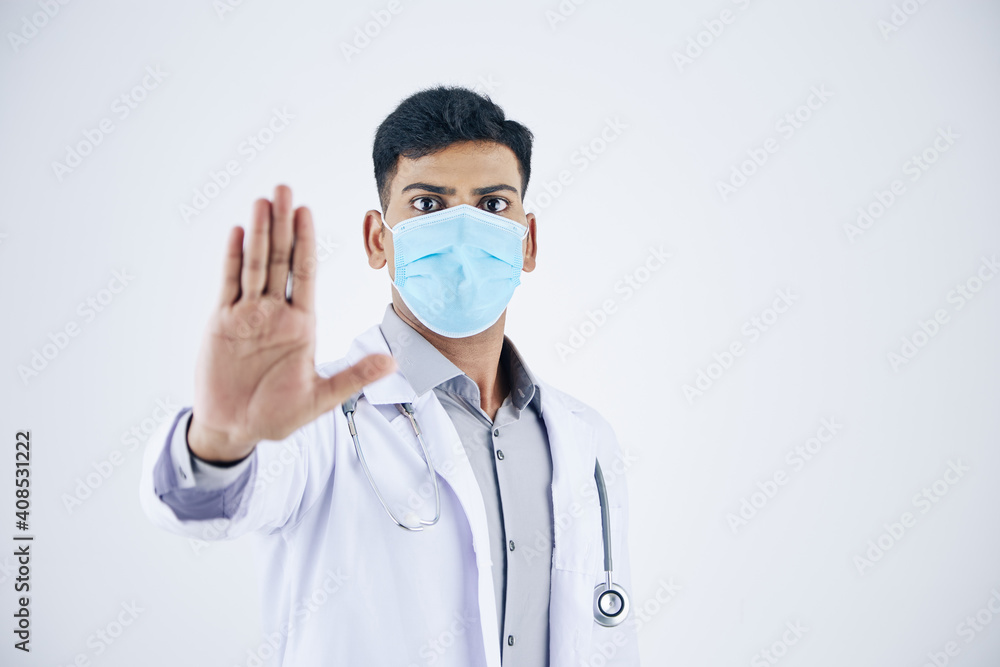 Serious young Indian general practitioner in medical mask making stop gesture, isolated on white Serious young Indian general practitioner in medical mask making stop gesture, isolated on white