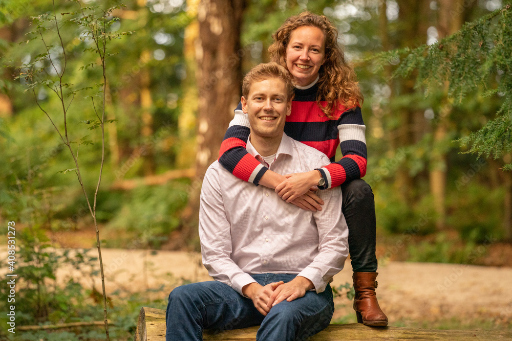 Fototapeta premium Beautiful enamored young couple sitting on a tree trunk in the forest. Facial expression is in love. In half body, woman behind the man