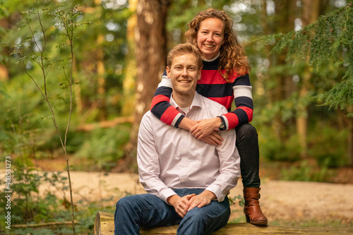 Beautiful enamored young couple sitting on a tree trunk in the forest. Facial expression is in love. In half body, woman behind the man