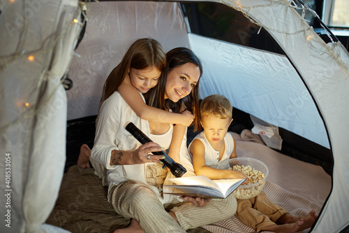 Mother reads a book of fairy tales for her children while sitting in a tent at night. Mom son and daughter reading a book with a flashlight in their hands © angel_nt