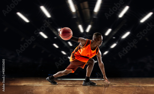 Team supporting. African-american young basketball player in action and motion in flashlights over dark gym background. Concept of sport, movement, energy and dynamic, healthy lifestyle. Arena's © master1305