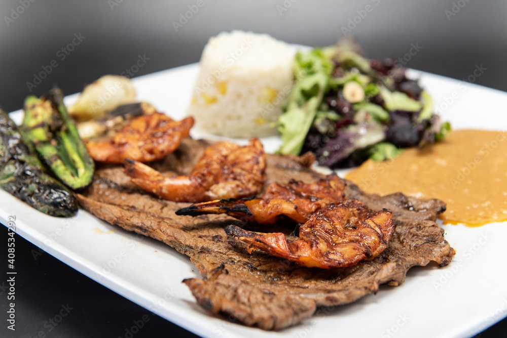 Grilled carne asada steak topped with split grilled shrimp served on a plate with rice and beans.
Grilled carne asada steak topped with split grilled shrimp served on a plate with rice and beans.