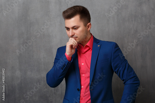 A handsome business man in a blue jacket and a bright pink shirt stands in thought with his hand to his chin on a gray background.