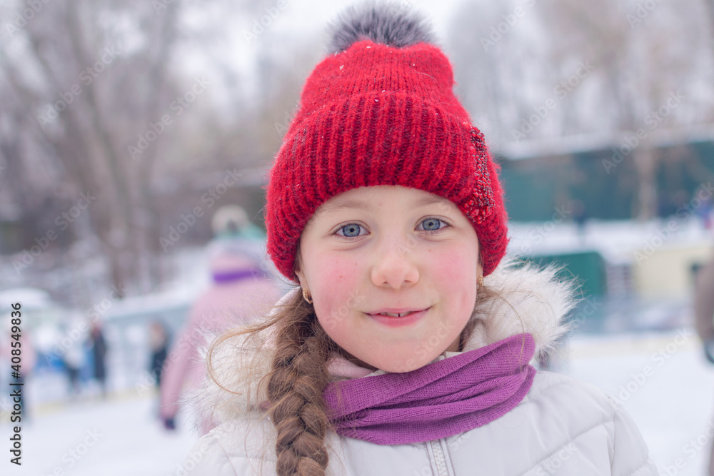 Winter portrait of a kid close up. Cheerful child in a red cap is smiling cute. Beautiful little girl is on the ice rink in the park Winter portrait of a kid close up. Cheerful child in a red cap is smiling cute. Beautiful little girl is on the ice rink in the park