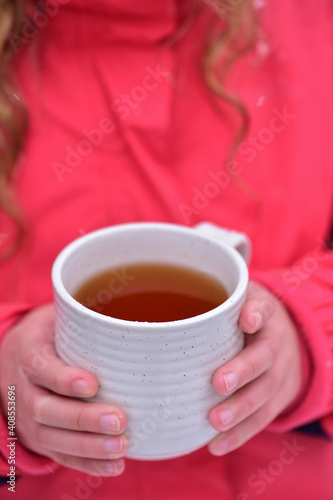 A white cup with tea in the hands of a girl in a pink jacket.