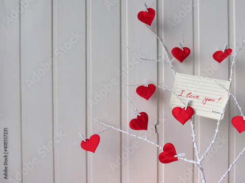 Red hearts on a white twig on a white wooden background. On the branch there is a note 