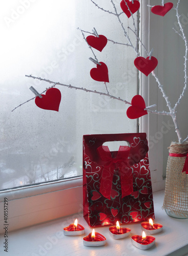  Gift in red decoration for Valentine's Day. Near a white twig with red hearts and candles on the background of the window