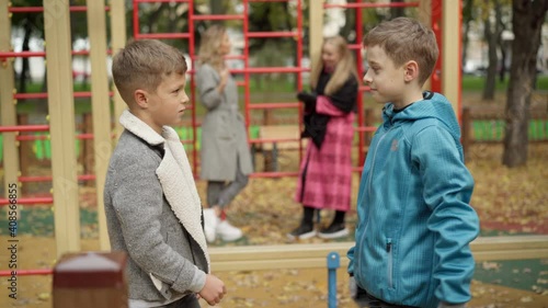Side view of Caucasian boys playing rock-paper-scissors and hide-and-seek with blurred relaxed women talking at background. Portrait of cheerful children having fun on playground on autumn day