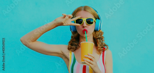 Portrait of modern young woman drinking a juice with headphones listening to music posing on a blue background