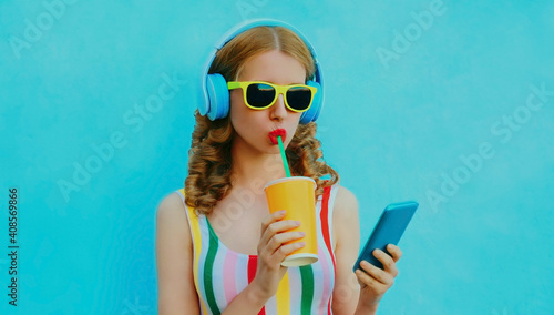 Portrait of modern young woman drinking a juice with headphones listening to music on a phone on a blue background