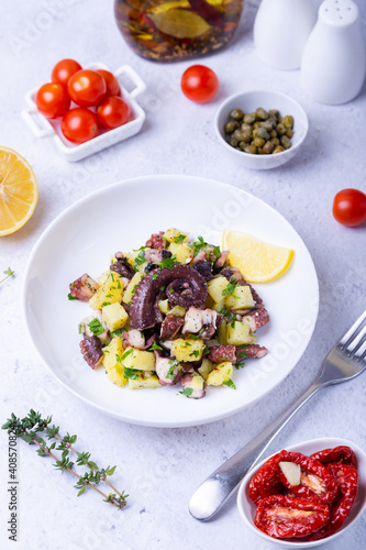Warm salad with octopus, potatoes, tomatoes, capers and lemon on a white plate. Close-up, white background.