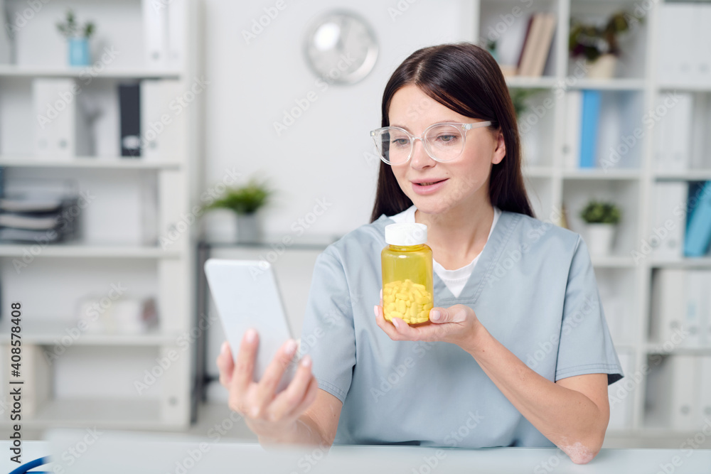 Young smiling female doctor showing bottle with new medicine to online patient Young smiling female doctor showing bottle with new medicine to online patient