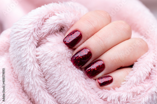 Female hand with beautiful manicure - dark red glittered nails on pink fluffy fabric, textile background with copy space. Selective focus
