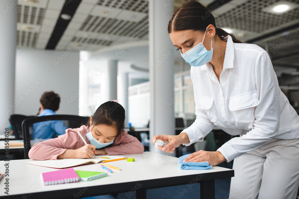 Fototapeta premium Teacher in mask cleaning table with antibacterial sanitizer and napkin
