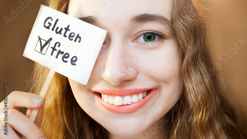 Cute girl holding a gluten free sign. Smiles and looks into the camera, covering one eye with a sign.