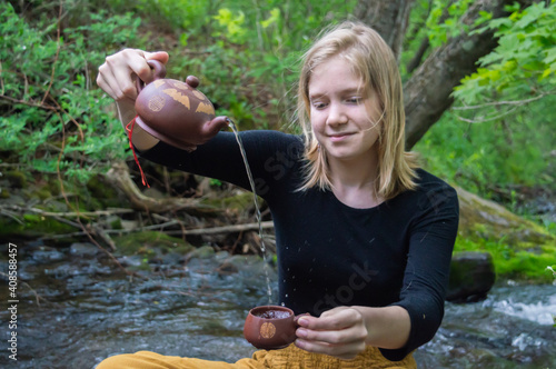 Portrait of a young smiling woman sitting cross legged on a rock on the shore of a river in the woodland, pouring tea or water in a tea cup. photo