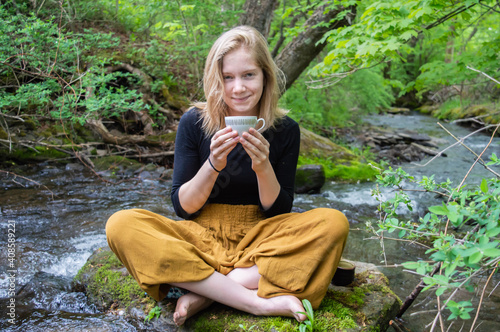 Portrait of a pretty young girl sitting cross legged on a rock on the shore of a river, holding a china tea cup with both hands and smiling at camera.