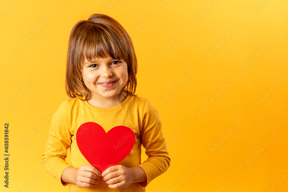 A cute baby girl on a yellow background in a yellow shirt holds a red heart. Concept Valentine's Day, love, smiley face, emotions A cute baby girl on a yellow background in a yellow shirt holds a red heart. Concept Valentine's Day, love, smiley face, emotions