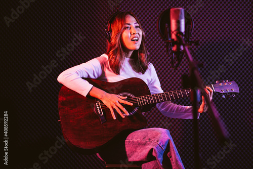 Bild auf Leinwand Pretty Asian female singer recording songs by using a studio microphone and pop shield on mic while playing guitar and wearing headphones in the studio