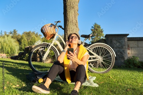 Happy mature woman with headphones sitting in the park, singing and listening music
