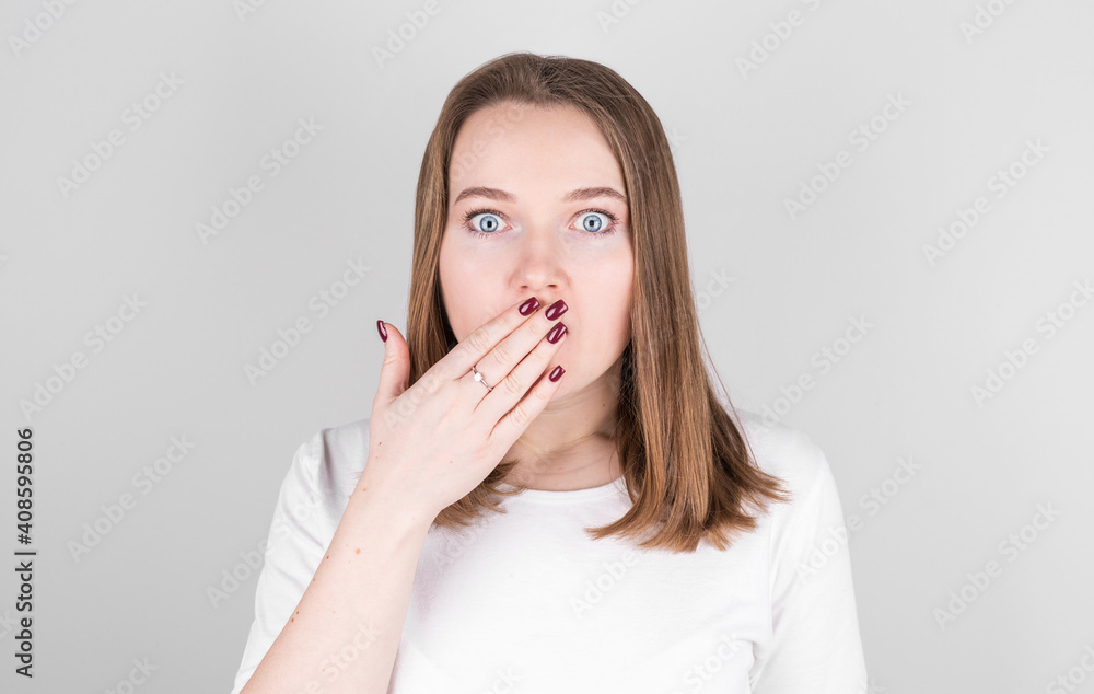 Нoung woman in a white T-shirt stands against a gray wall and covers her mouth with her hands in great surprise.