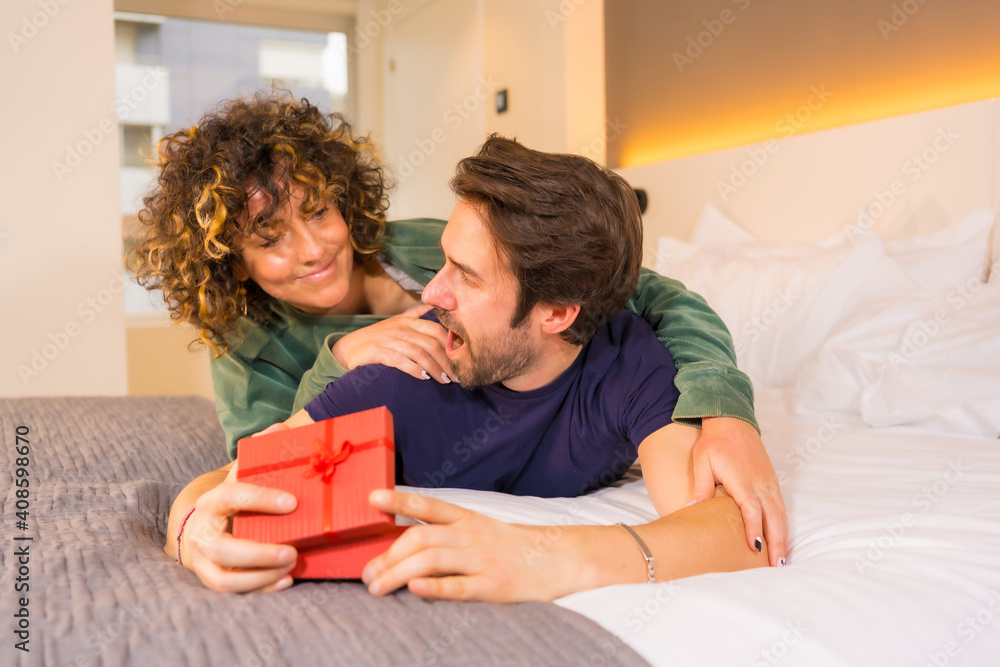 Valentine's Day, a young Caucasian couple in pajamas on the bed, the bride giving a nice surprise with a gift to the boyfriend Valentine's Day, a young Caucasian couple in pajamas on the bed, the bride giving a nice surprise with a gift to the boyfriend