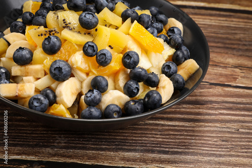 Fruit salad with blueberries, banana, orange, apple and kiwi on a black plate and wooden table. Vitamin cocktail. Summer taste.