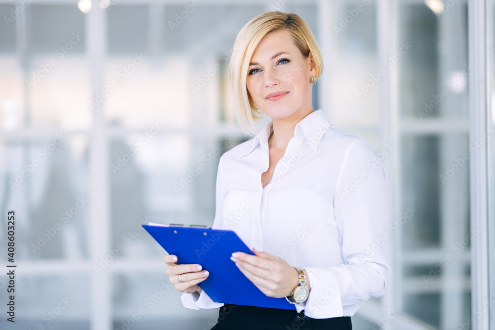 Beautiful blonde business woman in a white shirt with a folder of documents in an office building. Beautiful blonde business woman in a white shirt with a folder of documents in an office building.