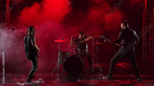 Camera slides over a group of rock musicians who play their instruments with crazy emotions in a dark studio illuminated by red lights. Drum set, electric and bass guitar. Slow motion.