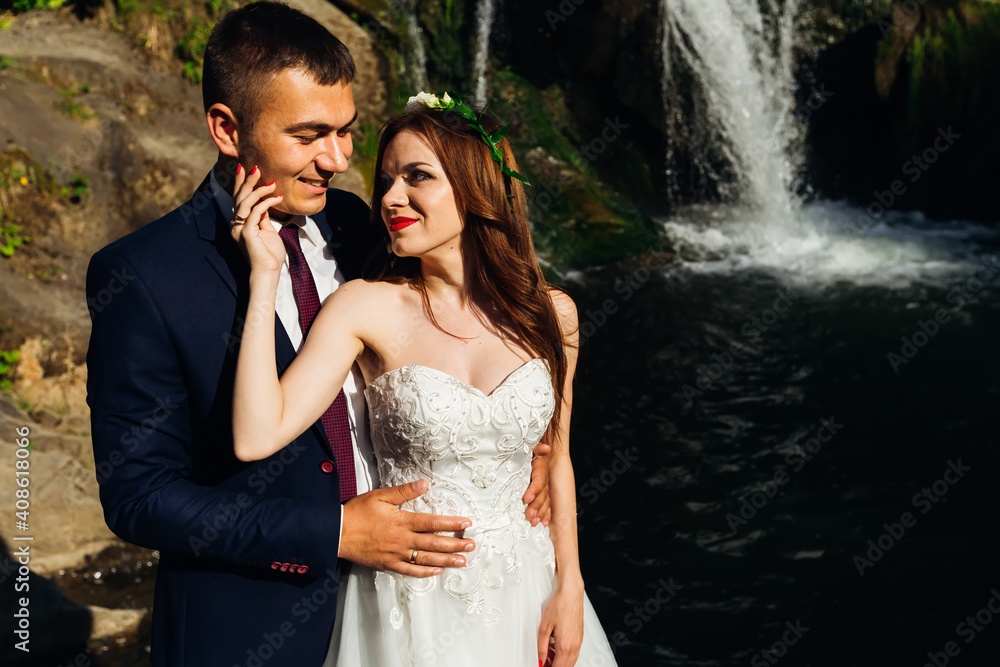 A young groom in a black dress coat with a red tie and a beautiful bride in a white dress are hugging against the backdrop of a cliff, rocks and a river.