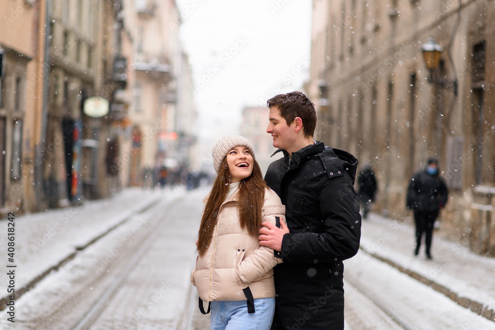 Having fun together at a christmas fairy with snowfall. Young cheerful couple is having a walk, enjoying, dressed warm, looking at each other and laugh, snowflakes all around.